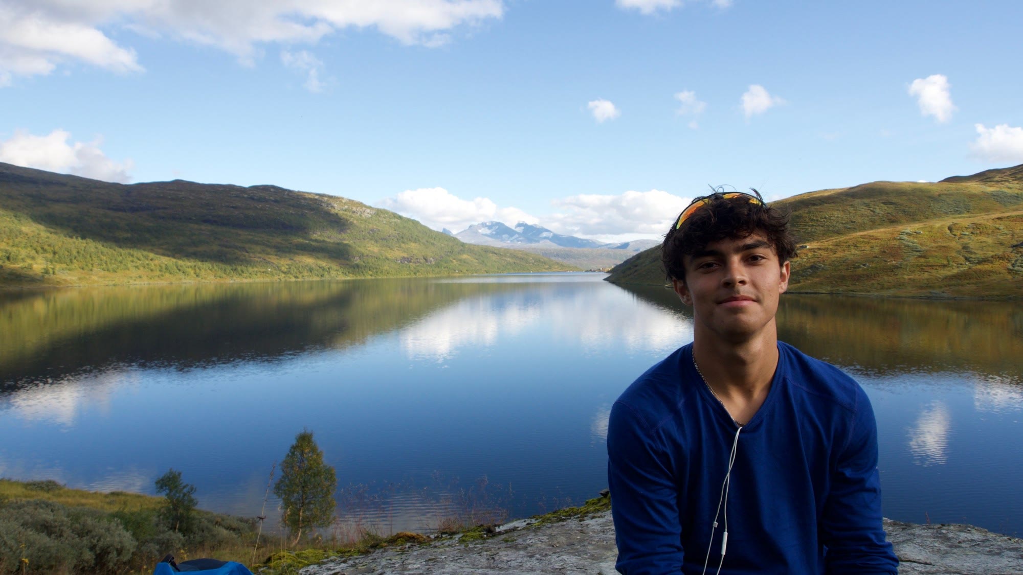 Joe sitting beside a still mountain lake, with green hills and snow-capped peaks behind